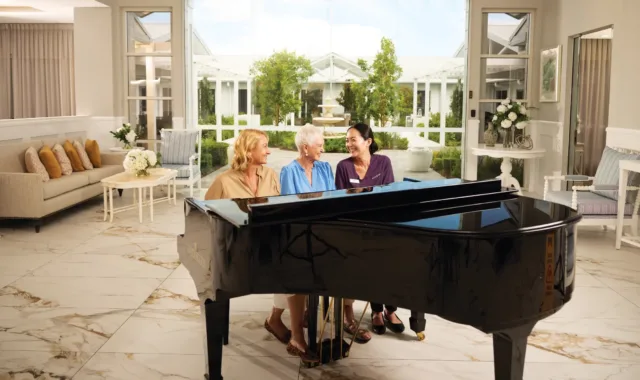Three women sit at a grand piano in a bright, elegant room with large windows overlooking a garden.
